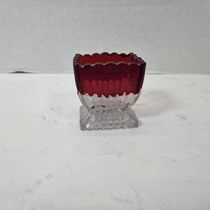 May include: A square glass vessel featuring a deep red upper section with a scalloped rim and a clear, textured base. The glass displays vertical ribbing along the sides, transitioning from the colored top to the transparent, footed bottom.