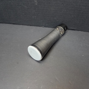 Könnte beinhalten: Eine schwarz-graue Taschenlampe mit weißer Endkappe. Die Taschenlampe hat einen strukturierten Griff und einen schwarzen Deckel mit einem kleinen Licht. Die Taschenlampe ist nach rechts abgewinkelt.