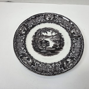 May include: A round, white and black decorative plate. The plate features a detailed landscape scene in the center, surrounded by an ornate border. The plate's design includes trees, buildings, and decorative elements.