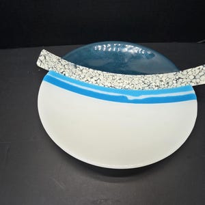 May include: A decorative art glass bowl with a white, blue, and speckled design. The bowl has a white base with a blue stripe and a teal-colored top. A speckled, rectangular piece sits across the top.