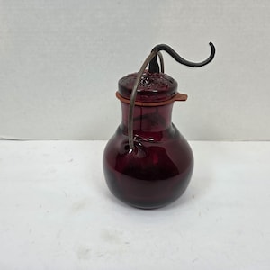 May include: A deep red glass bottle with a rounded body and a decorative lid. It features a metal handle and a brown leather or rubber ring around the neck. The bottle is likely for decorative or storage purposes.