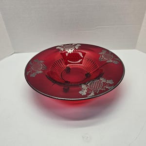 May include: A red glass bowl with a silver etched design of grapes and leaves. The bowl has a fluted edge and a raised center.