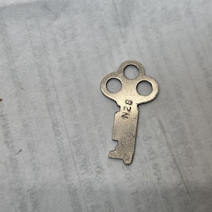 May include: A vintage metal key with a three-hole bow and a rectangular bit. The key is silver-colored and has the number "N28" stamped on it. The key is lying on a textured, off-white surface.