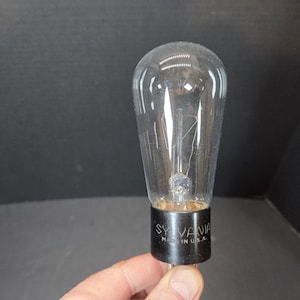 Puede incluir: Una bombilla de vidrio transparente con la parte superior redondeada y una base negra. La base tiene el texto "SYLVANIA MADE IN U.S.A.". La bombilla está sujeta por una mano.