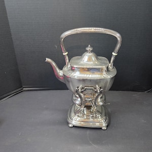 May include: A silver-toned metal tea kettle with a square-shaped body, a curved spout, and a high arched handle. The kettle rests on a matching square base with four small feet and features a central lid with a small knob on top.