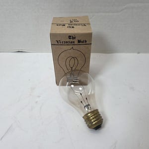 Op de afbeelding: Een heldere glazen gloeilamp met een messing voet, naast een bruine kartonnen doos. De doos is gemarkeerd met "The Victorian Bulb" met een lijntekening van een gloeilamp. De lamp is ongeveer 10 cm hoog.