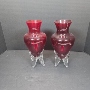 May include: Two ruby red glass vases with a flared top and a clear, three-legged base. The vases have a bulbous shape and are displayed against a dark background. These decorative vases are suitable for floral arrangements or as standalone decor.