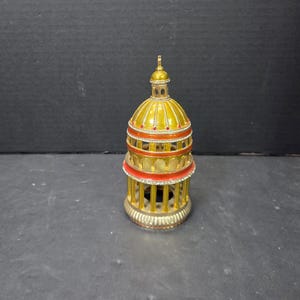 May include: A decorative, gold-colored miniature building with a domed roof. The structure features red and white accents, with a cage-like lower section. The overall design is ornate and detailed, suggesting a collectible item.