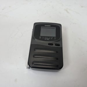 May include: A black Casio TV-100 portable television with a small screen, antenna, and channel selection buttons. The device has a rectangular shape with a ridged lower section and the Casio logo at the top.