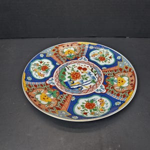 May include: Decorative ceramic plate with a detailed floral and bird design. The plate features a central oval design surrounded by six smaller floral designs. The color palette includes red, blue, yellow, and green accents.