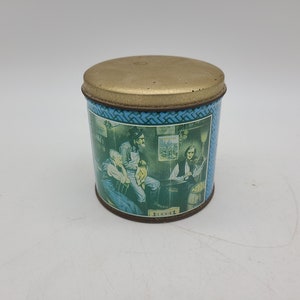 May include: A vintage blue and gold tin with a picture of three people sitting around a table. The tin has a gold lid and a blue and white patterned border.