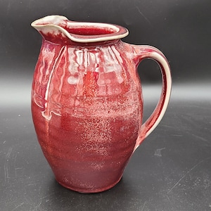 May include: A red ceramic pitcher with a white rim and handle. The pitcher has a slightly rounded shape and a textured surface.