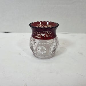 May include: A small glass vessel featuring a clear, faceted base with circular indentations and a deep red, scalloped rim. The red section is decorated with white, hand-painted swirling vine patterns and small leaf motifs encircling the top.