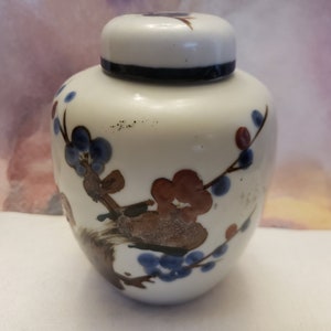 Könnte beinhalten: Eine weiße Keramikdose mit Deckel, verziert mit einem Blumendesign in Blau-, Rot- und Brauntönen. Die Dose hat einen traditionellen asiatischen Stil.