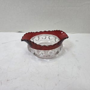 Puede incluir: Un cuenco de cristal transparente con un borde ondulado de color rojo intenso y un patrón exterior texturizado de hendiduras circulares. El cuenco tiene una parte superior ancha y abierta y una base redondeada, destacando el contraste de color.