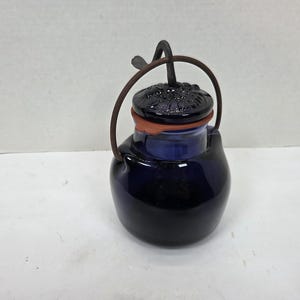 May include: A dark blue ceramic jar with a lid and a handle. The lid is dark blue with a floral design. The jar has a brown handle and a red-orange rim. The jar is sitting on a white surface.
