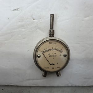 May include: Vintage voltmeter with a circular face and a needle pointing to the left. The meter has a silver-colored metal casing and a black needle. The face displays numbers and the words "Volts" and "Amps".