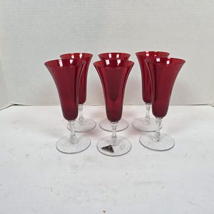 May include: Six red champagne flutes with clear stems and bases are arranged on a white surface. The flutes have a tulip shape and a vibrant red color. The bases are round and clear, providing stability.