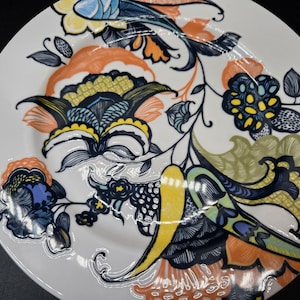 May include: A white ceramic dinner plate with a colorful floral design. The design features blue, green, yellow, and orange flowers with intricate details.