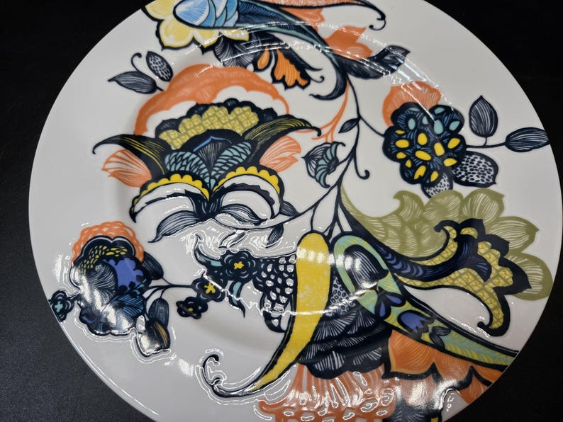 May include: A white ceramic dinner plate with a colorful floral design. The design features blue, green, yellow, and orange flowers with intricate details.
