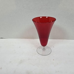May include: A ruby red glass champagne flute with a clear, circular base. The glass has a flared, conical bowl and a slender stem. The glass is approximately 9 inches tall.