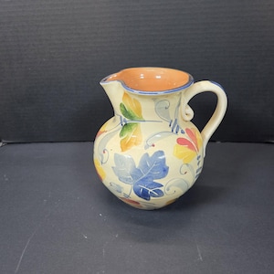 May include: A ceramic pitcher with a cream-colored base, adorned with colorful leaf patterns in blue, yellow, red, and green. The interior is orange, and the rim is outlined in blue. It has a curved handle.