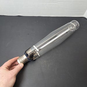 Puede incluir: Una bombilla transparente y alargada con una base plateada y componentes internos. La bombilla es sostenida por una mano sobre un fondo oscuro. El diseño de la bombilla incluye un elemento blanco central y soportes metálicos.