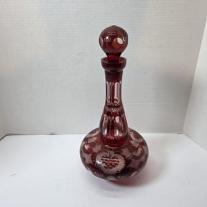 May include: A ruby red glass decanter with a matching stopper. The decanter has a bulbous base and a long, slender neck. The surface is etched with floral and geometric patterns. The stopper is round with a decorative design.