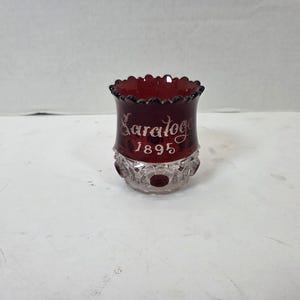 May include: A small, deep red glass tumbler with a scalloped rim. The upper portion features the etched white text "Saratoga 1895," while the lower section displays a clear, faceted pattern with circular indentations around the base.