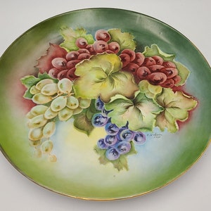 May include: A vintage porcelain plate with a hand-painted design of grapes and leaves. The plate has a green background with a gold rim and is signed "F.J. Alden 1934".
