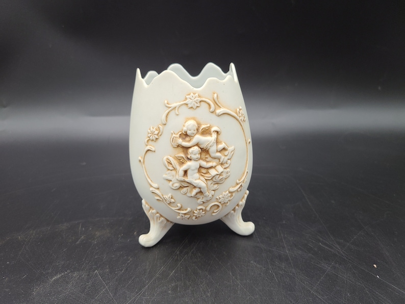 Puede incluir: Jarr&oacute;n de cer&aacute;mica blanca con forma de huevo con un dise&ntilde;o en relieve de dos querubines y motivos florales. El jarr&oacute;n tiene tres patas y un borde festoneado.