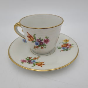 May include: A white teacup and saucer set with a gold rim and handle. The cup and saucer are decorated with colorful floral patterns, including red, purple, and blue flowers. The set is designed for serving tea.