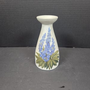 May include: A white ceramic vase with a flared top and a slender body. The vase is decorated with a blue floral design, featuring a cluster of blue flowers and green leaves. The vase is approximately 6 inches tall.