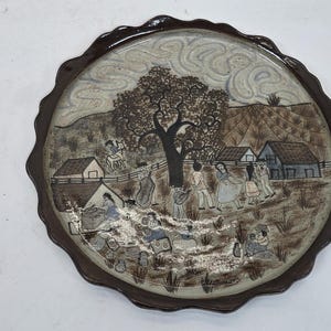 May include: A round ceramic plate with a brown rim and a detailed scene painted on the surface. The scene depicts a village with people, houses, and a large tree. The colors are muted, with browns, grays, and blues.