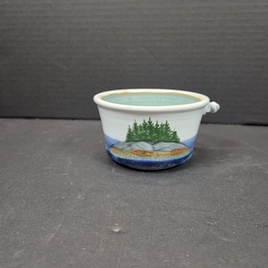 May include: A white ceramic bowl with a small handle, featuring a landscape design. The bowl has a light blue interior and a painted scene of green trees, a rock formation, and blue water. The base is dark blue.