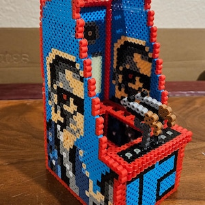 May include: A blue and red pixel art arcade game with a pixelated image of a person wearing sunglasses and a blue shirt. The game has a brown gun and a black and white control panel.