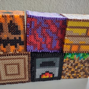 Può includere: Sei piccoli blocchi quadrati pixelati, ognuno con un diverso design di Minecraft. I blocchi sono impilati in due file di tre. La fila superiore presenta una zucca, un blocco rosso e viola e un blocco di legno. La fila inferiore presenta un blocco di legno, una fornace e un blocco di terra.