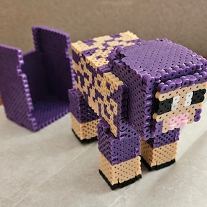 May include: A three-dimensional sheep figure constructed from purple and beige plastic fuse beads. The sheep has a blocky, pixelated appearance with black eyes and a pink nose. A separate, matching purple and beige bead structure sits nearby.