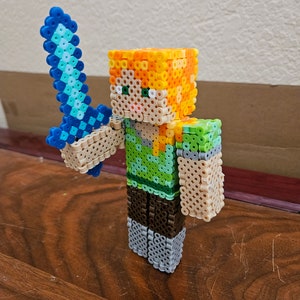 May include: A pixelated figure of a Minecraft character holding a blue sword. The character has orange hair, a green shirt, brown pants, and gray shoes. The figure is made of small plastic beads.