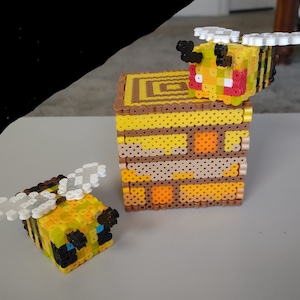 May include: Two yellow and black bee figures made of perler beads, one sitting on top of a yellow and brown beehive made of perler beads.