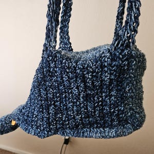 Puede incluir: Un bolso azul tejido a mano con una forma única. El bolso presenta un patrón de puntadas texturizadas y asas trenzadas resistentes. Un pequeño botón decorativo acentúa el lateral. El bolso está suspendido, mostrando su diseño.