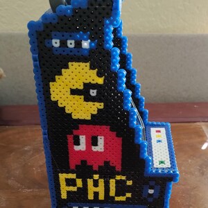 Peut inclure: Un mod&egrave;le de borne d'arcade en trois dimensions fabriqu&eacute; &agrave; partir de perles &agrave; repasser noires, bleues, jaunes et rouges. Le panneau lat&eacute;ral pr&eacute;sente un personnage Pac-Man jaune, un fant&ocirc;me rouge et le texte jaune PAC, avec des bordures en perles bleues.