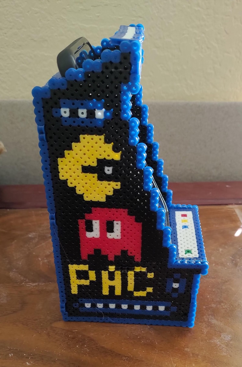Peut inclure: Un mod&egrave;le de borne d'arcade en trois dimensions fabriqu&eacute; &agrave; partir de perles &agrave; repasser noires, bleues, jaunes et rouges. Le panneau lat&eacute;ral pr&eacute;sente un personnage Pac-Man jaune, un fant&ocirc;me rouge et le texte jaune PAC, avec des bordures en perles bleues.