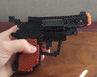 Peut inclure: Un revolver jouet fabriqué à partir de perles à repasser en plastique noir et marron. La poignée est marron, tandis que le cadre, le canon et le barillet sont noirs. L'extrémité du canon présente un accent en perle orange vif.