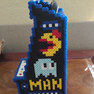 Peut inclure: Une vue lat&eacute;rale d'un petit support de borne d'arcade fabriqu&eacute; &agrave; partir de perles &agrave; repasser en plastique bleu, noir, jaune, rouge et bleu clair. Le design pr&eacute;sente un personnage jaune, un fant&ocirc;me bleu clair et le texte jaune MAN sur fond noir.