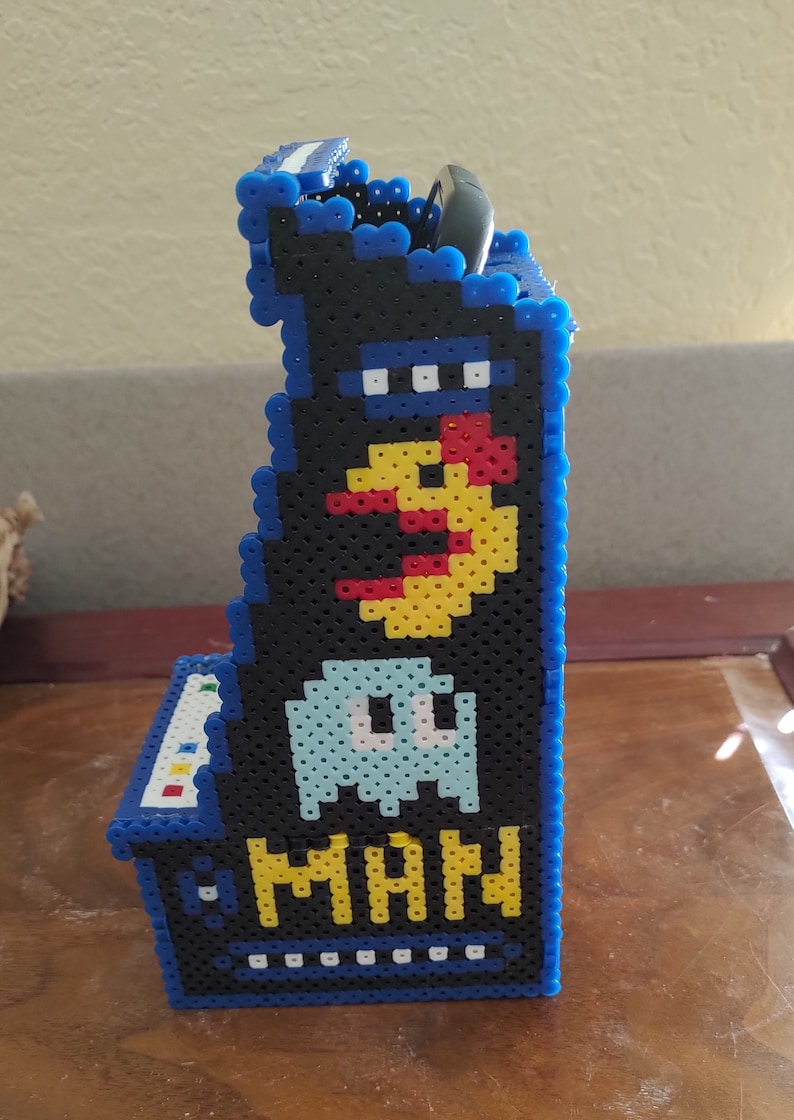 Peut inclure: Une vue lat&eacute;rale d'un petit support de borne d'arcade fabriqu&eacute; &agrave; partir de perles &agrave; repasser en plastique bleu, noir, jaune, rouge et bleu clair. Le design pr&eacute;sente un personnage jaune, un fant&ocirc;me bleu clair et le texte jaune MAN sur fond noir.
