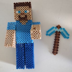 May include: A blocky character figure and a separate pickaxe tool constructed from small, fused plastic beads. The figure features a brown hair block, a tan face with blue eyes, a light blue shirt, and dark blue pants. The pickaxe is light blue and brown.