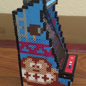May include: A pixel art phone stand made of colorful beads, featuring a retro arcade game design with a brown character and a blue background. The stand is designed to hold a smartphone in a vertical position.
