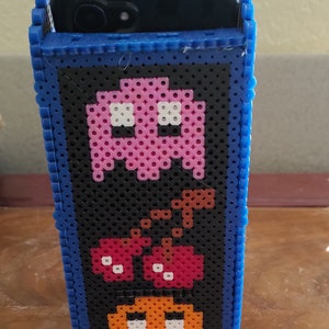 Peut inclure: Un support de t&eacute;l&eacute;phone rectangulaire fabriqu&eacute; &agrave; partir de perles &agrave; repasser en plastique bleu, noir, rose, rouge et orange. La face avant pr&eacute;sente un motif pix&eacute;lis&eacute; d'un fant&ocirc;me rose, de deux cerises rouges et d'un fant&ocirc;me orange sur fond noir.