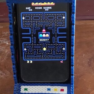 Peut inclure: Un support de smartphone style borne d'arcade en perles &agrave; repasser bleues. Le cadre pr&eacute;sente une barre sup&eacute;rieure blanche avec des accents rouges et une base orn&eacute;e d'un motif de fraise pix&eacute;lis&eacute;. Un smartphone est ins&eacute;r&eacute; affichant un jeu Pac-Man.